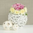 On a clean white tabletop, a carefully arranged rose centerpiece creates a soft, romantic focal point that feels ideal for a wedding breakfast in one of Oxford's historic colleges or a chic celebration near the Botanic Garden. The flowers form a compact, rounded bouquet, with each rose head nestled tightly against its neighbour, so the viewer sees a dense cushion of blooms rather than individual stems. The colour story is gentle yet distinct: blush and lavender-pink roses meet creamy yellow and ivory roses, their tones blending where they touch and creating a subtle swirl of pastel hues. Woven between the roses, fine sprays of white baby's breath lend a light, airy texture, like small clouds drifting among the petals. Supporting this floral crown is a distinctive spherical vase, fashioned from slim, white-painted branches or wicker that have been intertwined into an open, cage-like globe. Its intricate lattice catches the light and casts delicate shadows, offering a contemporary take on rustic design. To the left, two smaller decorative spheres made from pale rattan or natural wood echo the woven pattern of the vase, one slightly larger than the other, adding depth and visual rhythm to the scene. The entire arrangement is set against a softly textured beige or light taupe background, similar to fabric or linen, which keeps the focus firmly on the roses while maintaining a cosy, welcoming atmosphere. The overall impression is of fresh, hand-crafted beauty and quiet refinement, a centerpiece that would sit comfortably on tables from Summertown townhouses to riverside venues along the Thames in Oxford.