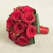 An exquisite hand-tied bouquet of deep crimson roses sits upon a soft neutral cloth, evoking the serene sophistication of Oxford's historic surroundings. Each of the eight to ten roses glows with a classic ruby hue, petals gently unfurled to reveal velvety layers and generous depth. Dotted among them are glossy red hypericum berries, introducing a lively texture and pop of festive colour-similar to those glimpsed at local Oxford markets. A border of broad, deep green leaves wraps neatly at the base, creating natural structure and drawing the eye toward the central blooms. The stems are skillfully bound with a shimmering red satin ribbon whose finish is smooth, secure, and undeniably refined. Lighting is soft and even, delicately accentuating each petal and the subtle shine of both the ribbon and berries. The overall mood is one of romantic celebration-a design suitable for weddings in the Oxford colleges, anniversary surprises, or meaningful declarations of love. This bouquet, available for prompt local delivery, brings with it the character and tradition of Oxford, expertly assembled with attention to every luscious detail.