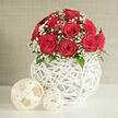 At the heart of this image is a lush dome of red roses, gathered so closely that their petals almost touch, forming a rich, velvety canopy of colour. Each bloom is at full, luxurious openness, the deep ruby tones recalling classic romance and the kind of timeless elegance many couples in Oxford choose for intimate wedding celebrations. Threaded delicately between the roses, sprays of white baby's breath drift like tiny clouds, their airy texture and bright whiteness offering a gentle, lacy frame that softens the intensity of the crimson. A few dark green leaves appear at the edges of the bouquet, grounding the arrangement in natural freshness, as though the flowers have just been conditioned in a local florist's studio near Jericho or Summertown. Supporting this floral crown is a sculptural white vessel fashioned from interwoven strands, creating a hollow spherical vase with a crisscross pattern reminiscent of wicker or rattan. The open lattice casts subtle shadows across the clean white surface on which it rests, adding depth and a whisper of movement. Nearby, two decorative orbs made from paler, cream-toned wicker lie casually on the tabletop, mirroring the vase's structure and contributing to an effortlessly curated look. The background is simple and neutral, perhaps a softly textured beige wall like those found in many Oxford townhouses, ensuring all attention remains on the interplay of reds, whites, and greens. The soft, balanced lighting enhances the serene mood, making this composition ideal for illustrating wedding flowers, engagement gifts, or romantic home décor suitable for same-day delivery across Oxford's historic colleges and riverside homes.