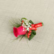 Presented on a subtle, light-beige linen surface, this boutonniere centres on a fierce magenta rose whose tightly furled core relaxes into soft, velvety petals, suggesting a bloom just beginning to reveal itself. Surrounding it, sprays of pristine white gypsophila create a mist-like halo that softens the silhouette and brings a whisper of old-fashioned charm. Scattered among those tiny white blooms, a few bright red hypericum berries add playful punctuation-small, glossy beads that catch the light and mirror the rose's intensity. Wispy, grass-like foliage fans out with an airy, natural movement, while broader, folded green leaves are tucked in to build a sculpted frame and visual weight. The base is bound with a rich crimson satin ribbon, wound tightly and finished in a small, elegant bow whose sheen contrasts beautifully with the linen's muted texture. The whole arrangement lies diagonally in the frame, inviting the eye to travel from bloom to ribbon and notice the careful layering and balance of textures. In a quiet Oxford morning light the colours feel both bold and tender; the scent is a soft, green-rose note that doesn't overwhelm. Perfect for a groom, groomsman, or a guest after a ceremony in Jericho or a celebratory meal near Summertown, this boutonniere reads as intimate and joyful, handcrafted with the kind of attention to detail local customers expect. Available fresh and ready for same-day delivery across Oxford when needed for that special day.