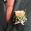 This boutonniere is composed with the exacting eye of a neighbourhood florist: a single peach rose, immaculate and softly luminous, rests against a charcoal suit lapel, its coral-pink petals curling inward like the first blush of morning in a quiet Oxford lane. The rose's surface is velvet-smooth, the petal edges kissed with a creamy blush that gives depth and warmth when caught by ambient light. Surrounding the central bloom, sprays of tiny white baby's breath - gypsophila - form a delicate halo, their cloud-like clusters softening the silhouette and lending an ethereal, celebratory quality reminiscent of confetti at a corner of Gloucester Green. Slender green foliage provides both contrast and structure; a gently curved leaf props the rose while wisps of fern add a hint of wildness, suggesting hedgerows along Botley Road after a light rain. A narrow satin ribbon, matched to the tie's coral hue, secures the stems in a tidy bow, the satin's sheen mirroring the tie and drawing the eye to the boutonniere's craftsmanship and restraint. The charcoal fabric beneath acts as a muted backdrop, amplifying the peach tones and underscoring the boutonniere's suitability for an elegant wedding or a thoughtful anniversary celebration. There is a faint, uplifting scent from the rose - soft, floral, and promising - that completes the sensory impression. This little arrangement feels personal and assured, the sort of finishing touch you might choose for a groom or dear guest walking from Jericho toward the university chapels on a sunlit afternoon.