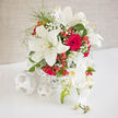 A lush wedding arrangement of snowy lilies, classic red roses, soft orchids, and berries sits composed and harmonious, like a centrepiece set for a reception in an Oxford hotel near Broad Street. Large ivory lilies open dramatically, each bloom a graceful star with slightly ruffled edges, their smooth petals catching the light and reflecting a feeling of new beginnings. Nestled alongside, deep red roses, tightly layered and velvety, provide contrast and depth, their colour reminiscent of old leather-bound books in the Bodleian Library. Gently cascading from the design, pale orchid blooms with creamy white petals and subtle blush centres drape over the edge, lending movement and an almost silk-like texture, as though the flowers themselves are leaning into the celebration. Wisps of gypsophila weave through the arrangement, softening the transition between the bold reds and crisp whites, creating a cloud-like veil that feels both romantic and modern. Small clusters of shiny red berries punctuate the bouquet, echoing the joyful energy of wedding guests spilling out into Radcliffe Square after a ceremony. The rounded, carefully balanced shape makes it easy to imagine this arrangement gracing a head table, or placed near the entrance to welcome guests to an Oxfordshire barn wedding. Gentle greenery peeks out from beneath the focal blooms, adding structure without overpowering them. The overall impression is refined yet heartfelt, like a love story grounded in tradition but open to contemporary touches. Every bloom seems placed with intention, reflecting the practiced eye of a local florist attuned to the tastes of couples marrying across Oxford and the nearby villages.