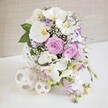 A beautifully composed floral centerpiece sits on a clean white tabletop, the kind you might see dressing a wedding table in an Oxford restaurant near the Covered Market or in a light-filled North Oxford home. At its core, several immaculate white Phalaenopsis orchids unfold, their smooth, wide petals catching the light with a satin sheen, each bloom revealing a delicate golden-yellow and soft orange center that brings warmth and depth. Encircling the orchids, generous clusters of roses in blush pink, lilac, and pastel lavender open luxuriously, layers of velvety petals spiralling outward to create a rich, romantic texture. Among them, smaller ruffled flowers-likely lisianthus or Eustoma-show off white petals finely edged in purple, adding a subtle, painterly accent. Feather-light sprays of white baby's breath are threaded throughout, making the bouquet appear airy and almost cloud-like, while trailing stems of light purple flowers cascade gracefully over the rim, giving the design a soft, flowing line. Glossy green leaves and finer, fern-like foliage peek out between the blooms, grounding the pastel tones with fresh, natural contrast. The arrangement is cradled in an eye-catching spherical vase crafted from white woven strands, its open lattice resembling delicate rattan or yarn wrapped into an intricate ball, allowing glimpses of shadow and negative space. On the same white surface, two decorative woven spheres in a cream or natural shade sit to one side, one slightly larger than the other, echoing the vase's texture and reinforcing the modern yet gentle aesthetic. Behind, a softly textured beige wall remains unobtrusive, letting the colours and craftsmanship take center stage. The overall impression is one of serene celebration and refined taste, perfect for marking a wedding, anniversary, or special gathering in Oxford's historic heart.