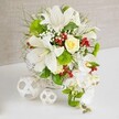 An elegant white and green wedding-style table arrangement sits poised on a crisp white surface, evoking the calm atmosphere of a celebration in an Oxford riverside restaurant or college dining hall. The flowers emerge from a round white woven wicker basket whose intricate pattern adds discreet texture and a handmade feel. At the heart of the display, several large white lilies open generously, their trumpet-shaped blooms showing fine dark speckles and warm yellow stamens, suggesting a gentle, perfumed presence in the room. Above and between them, a single pale cream-yellow rose holds pride of place, its tightly layered petals forming a soft focal point amid the surrounding white. Flowing down from the right side, a cascade of white phalaenopsis orchids drapes elegantly, each bloom smooth and luminous, giving the arrangement a waterfall or teardrop silhouette that suits bridal and formal settings. Around these statement flowers, bright green pom-pom chrysanthemum blooms add rounded, cushiony shapes, enhanced by wispy, feathery foliage and slender leaves that bring movement and depth. Tiny sprays of white baby's breath appear like fine confetti scattered throughout, while clusters of rich ruby-red berries punctuate the design with glowing, jewel-like accents. To the left on the table, two white wicker spheres, one small and one larger, echo the basket's weave and create a balanced, sculptural vignette. The backdrop is a soft beige fabric, gently textured and unobtrusive, allowing every petal, leaf, and berry to stand out clearly. The composition feels serene, luxurious, and joyfully celebratory, ideal as a wedding table centerpiece or an impressive gift delivered across Oxford for a special occasion.