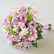 A vibrant Oxford bouquet brimming with fresh, romantic hues takes centre stage on a soft, neutral cloth-the perfect backdrop for its lively colours. At first glance, fully bloomed roses in shades of blush and lilac, their petals perfectly unfurled, create a sense of timeless elegance and charm, reminiscent of the walled gardens dotted throughout Oxfordshire. Dotted between these classic blooms are rounded clusters of bright chartreuse button chrysanthemums, their playful shapes and vivid colour recalling the energy of a summer's day in South Park. Trumpet-shaped Alstroemeria lilies, their pink petals etched with yellow and maroon, add lightness and movement, while delicate white gypsophila threads through every layer, softening the profile with a gentle, almost ethereal touch. Lush greenery frames the florals, anchoring the arrangement with nature's fullness. Each stem is gathered neatly and wrapped in soft, satiny pink ribbon, suggesting this bouquet was made to be held close-perhaps for a walk through the streets near Radcliffe Square or to accompany a moment of celebration at the Bodleian. Ideal for weddings, anniversaries, and bridal features, the attention to colour harmony and textural detail exemplifies the craftsmanship of Oxford's local florists, ensuring each bouquet is as memorable as the city itself.