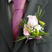 This boutonnière is captured as though resting against a tailored lapel, a finely balanced composition that mirrors the quiet sophistication of a wedding unfolding in central Oxford. At its core is a single tight pink rose, the petals smooth and luminous, blushing gently as though just beginning to open to the day. Around it, slender sprays of pine-like greenery and folded leaves create a frame of rich, cool green tones, reminiscent of shaded paths through University Parks. Nestled among these textures are clusters of white gypsophila, their tiny, cloud-like flowers drifting lightly around the rose and lending an airy sweetness that softens the whole design. Near the centre, a fresh green button chrysanthemum stands out with modern clarity, its circular form and clear lime colour giving the piece a crisp, contemporary edge-perfect for a groom walking from an Oxford college room along Broad Street toward the ceremony. The stems are gathered and wrapped with a subtle ribbon, the binding precise and tidy, hinting at the experienced hands that assembled it. Under gentle light, the contrasting greens, whites, and soft pinks appear both calm and quietly celebratory, like the hush before music starts in a chapel near St Giles'. There is an effortless grace in the way each element supports the others: the rose offering romance, the gypsophila whispering charm, the chrysanthemum suggesting freshness and intention. This small floral accent feels crafted for meaningful moments-a prom arrival near Cowley Road, perhaps, or a wedding reception in Summertown-where a single, thoughtful detail can say more than any words pinned beside it ever could.