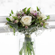 A softly glowing sympathy bouquet of white and green stands poised in a clear glass vase, illuminated by gentle, diffused light that could easily be streaming through a townhouse window off St Giles' in Oxford. Central to the arrangement are several large white roses, each bloom opening generously, their velvety petals forming elegant spirals with a creamy ivory depth at their centres that suggests tenderness and care. Surrounding these focal roses, sprays of gypsophila create a light, cloud-like frame, their tiny white blossoms so numerous and fine that they lend the bouquet a veil of softness, like distant fog along the Oxford Canal. Threaded between are stems of alstroemeria, some still in bud and others beginning to unfurl, displaying narrow petals tinged with lavender and pale purple, with discreet stripes that offer gentle visual interest. At the base, broad, polished deep-green leaves are wrapped around the stems in smooth bands, giving the arrangement structure and a sense of being gently held. Slimmer, bright green leaves wander upward through the bouquet, adding texture and freshness in a way that feels reminiscent of nearby university gardens in spring. The transparent cylindrical vase reveals the straight, vivid green stems, standing in clear water that reflects the soft light. A subtle natural tie gathers the stems just above the water, maintaining the classic, rounded form of a professionally hand-tied bouquet. The overall impression is one of quiet strength, purity, and solace, perfect for sending condolences or placing in a peaceful corner of an Oxford home as a thoughtful reminder of love and remembrance.