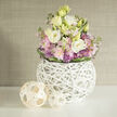 Nestled on a smooth white shelf, this compact yet generous bouquet pairs creamy white and pale buttery-yellow lisianthus with soft clusters of blush-pink Stock and delicate, spiky purple Allium blooms, all woven together with fresh green foliage that breathes life through the design. A ring of deeper green leaves hugs the base, subtly pulling the composition into a neat, rounded profile. The flowers are held in a sculptural spherical vase crafted from a web of slim white branches: an airy, handcrafted lattice that gives the arrangement lightness and allows stems to peek through, balancing form and transparency. Beside the vase, two natural rattan-like spheres in light wood tones rest as casual accents; their open weave mirrors the vase's texture and adds an organic rhythm to the still-life. The backdrop is a soft beige-greige wall with a whisper of texture, like grasscloth, which warms the scene and makes the pastel blooms feel fresh and tactile. The overall mood is calm and refined - a bouquet that would suit a foyer near High Street or a tranquil spot in a Summertown flat, offering a quiet, springlike fragrance and a gentle visual rest. Crafted with an eye for proportion and material, this arrangement suggests both modern interior styling and the comforting hand of traditional floristry, made to soften rooms and mark small domestic moments with subtle elegance.
