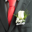In soft, directional light the ivory rose at the centre of this boutonnière becomes almost luminous against the charcoal jacket, the petals' velvety surface catching shadow and glow in equal measure. Sprays of baby's breath fan gently at the base, their tiny white blossoms creating a cloud-like counterpoint that gives the rose room to breathe. Fine slivers of fern and twisted leaves arch with rhythmic movement, lending depth and a slightly wild, natural line that contrasts the groomed restraint of the suit fabric. The dark green foliage frames the bloom, providing contrast and emphasising the rose's cool, creamy tone while a narrow white ribbon is tied in a modest bow, its soft sheen hinting at ceremony without ostentation. The tactile crispness of the flowers and the faint, refreshing scent of rose are almost palpable, conjuring the sensation of dew on early summer mornings in Oxford. As a local florist at Flowers Oxford, we often create this refined style for couples who exchange vows near Christ Church or stroll through University Parks for photographs; it complements both the grandeur of formal wear and the intimacy of close family moments. This boutonnière is deliberately understated-an emblem of loyalty and celebration that sits comfortably on a lapel, ready to be worn with calm confidence as the wearer moves through the day's important moments, quietly speaking of care and presence.