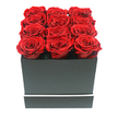 This photograph features a strikingly modern rose arrangement that exudes quiet luxury, as if it has just been set down on a polished table in a stylish apartment overlooking Oxford's Botley Road. At the centre of the composition is a square matte black gift box, its surfaces smooth and unadorned except for a thin, crisp white stripe circling the lower section like a minimalist frame. Nestled inside are twelve preserved red roses, arranged so precisely in a 3x4 grid that their open blooms form a continuous field of red, neatly filling the top of the box and rising slightly above the rim. Each rose is at full bloom, with layers of velvety petals spiralling inward to form rich, textured rosettes. The crimson hue is deep and saturated, with subtle hints of darker burgundy in the shadows of the petals and brighter ruby tones at their edges, creating a sense of depth and richness. The lighting is gentle and even, casting soft, understated shadows that define the edges of the petals and the corners of the box without any stark contrasts. A pure white background isolates the arrangement, allowing the interplay between black, white, and red to stand out clearly, much like the way bold colour accents enliven many contemporary Oxford interiors. The mood is romantic yet controlled, ideal for a milestone anniversary, a sophisticated Valentine's gift, or an elegant thank-you gesture to someone in Jericho, Summertown, or the city centre. The preserved nature of the roses suggests a message of enduring affection, their lasting beauty perfectly suited to premium flower delivery across Oxford when a long-term keepsake feels more meaningful than fleeting blooms.