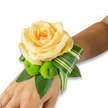 There is a gentle romance woven into this wrist corsage: a single, buttery yellow-tinged rose blooms at the centre, its layered petals unfurling in soft, luminous folds that catch the light like morning on the Isis. Tucked beneath the rose are two almost-spherical lime-green button chrysanthemums, their dense, spiky florets offering a playful, tactile contrast to the rose's velvety surface. A ribbon-like, variegated leaf with sharp white stripes curves artfully to the side, its folded architecture lending a modern sweep that balances the arrangement, while broad, glossy salal or ruscus leaves form a deep green backdrop and cradle the flowers. The corsage sits on the inner wrist and hand of a person, set against a clean white studio background so every subtle hue and texture reads clearly: the warm glow of the rose, the bright, cheerful green of the pom-poms, and the cool stripe of the variegated foliage. Although the fastening - an elastic band or narrow ribbon - is not visible, the design reads wearable and secure, made to move with a first dance or a soft embrace. The scent is implied: a faint trace of rose and freshly cut greenery, as if made on a summer morning at a shop near the Botanic Garden. Ideal for weddings, proms, or anniversary celebrations, this handmade wristlet radiates quiet anticipation and tactile intimacy. A handcrafted floral accent that feels both celebratory and intimately personal, it promises to be remembered in photographs and in the warm hush of close moments.